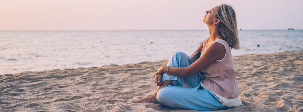 Mujer sentada con las piernas cruzadas en una playa al atardecer, mirando hacia arriba, con el océano al fondo.