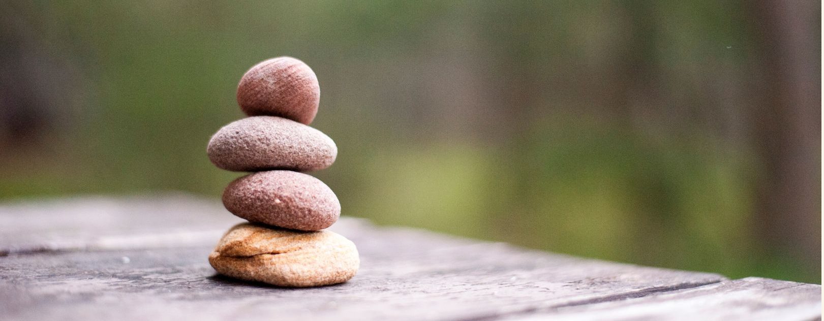 Una pila de cuatro piedras lisas en equilibrio sobre una superficie de madera, con un fondo de bosque verde borroso, que evoca la atención plena y la paz que a menudo se buscan en la meditación.